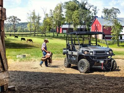 Person walking toward a 2018 Kawasaki Mule Pro-FXT™ Ranch Edition with a saddle and cows and a barn behind them